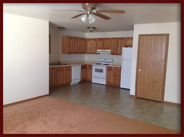 a kitchen area in the corner of a large room. The corner has cabinets, counters, a white dishwasher, white stove and white refrigerator. An interior door is on the same wall.
